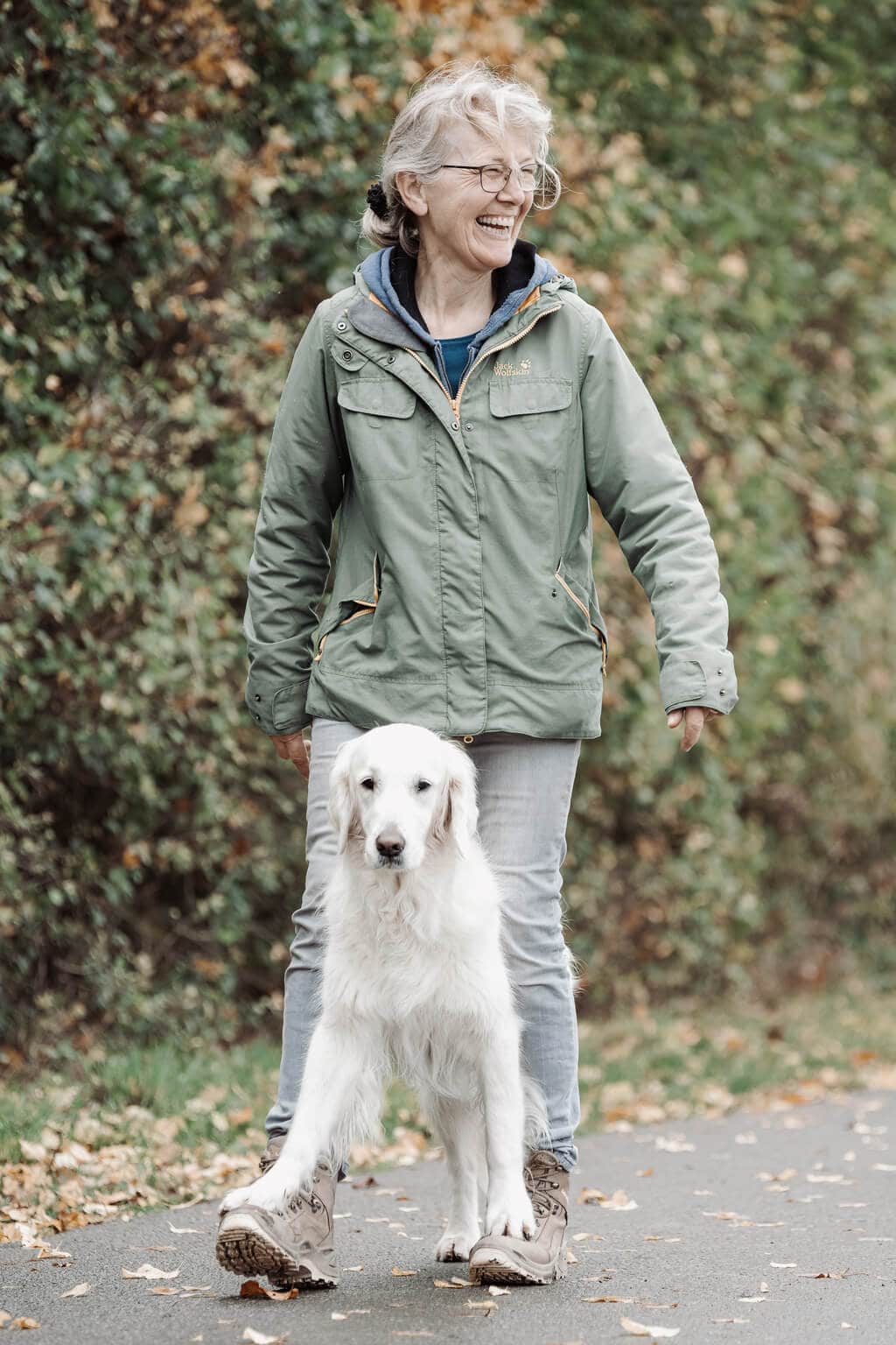 Ein weißer Hund steht zwischen den Beinen seiner Hundehalterin und hat seine Vorderpfoten auf ihren Füßen. Gemeinsam laufen sie über einen Weg.