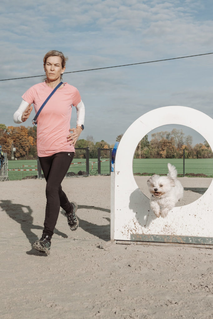 Eine Frau rennt über Sandboden daneben springt ein kleiner, weißer Hund durch einen Ring beim Turnierhundesport Kurs