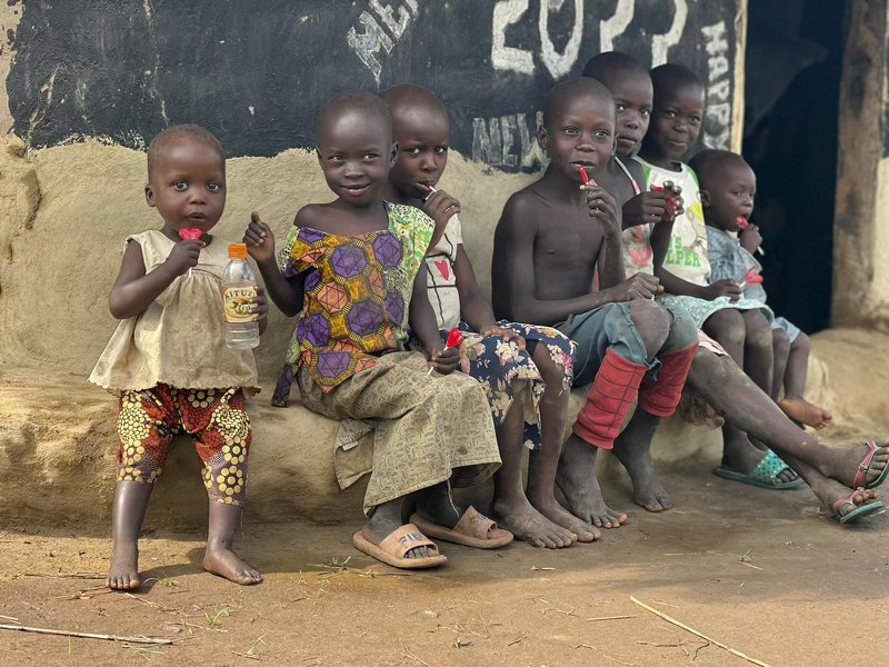 7 Kinder sitzen vor einer Hütte in Uganda und haben Lollypops in Form von roten Herzen in der Hand